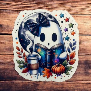 Könnte beinhalten: Ein weißer Geist mit einer schwarzen Schleife liest ein Buch mit einem Galaxie-Design. Der Geist hält eine Tasse Kaffee und es gibt einen Kürbis und Herbstblätter um ihn herum.