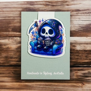 May include: A blue and white glitter bookmark featuring a cartoon Grim Reaper sitting in a blue armchair and reading a book that says "Always Reading Trash".