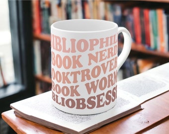 Puede incluir: Una taza de cerámica blanca con las palabras "Bibliophile, Book Nerd, Book Lover, Booktrovert, Book Worm y Bibliobsessed" en tonos rosas y marrones. La taza está sobre una superficie de madera con un libro abierto, con una estantería borrosa al fondo.