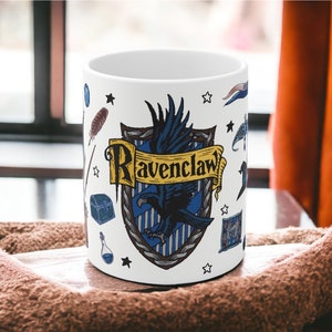 Puede incluir: Taza de cerámica blanca con un escudo de la casa Ravenclaw azul y amarillo de la serie Harry Potter. El escudo representa un cuervo y las palabras "Ravenclaw" en una pancarta amarilla. La taza está decorada con estrellas, una pluma, una botella de poción y otros símbolos mágicos.