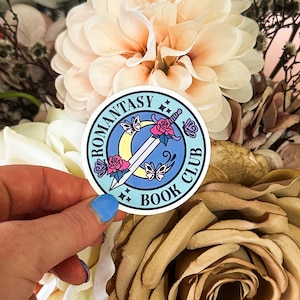 May include: A blue and white round sticker with the text "Romantasy Book Club" and a sword, moon, and butterflies.