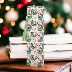 May include: A pink and green bookmark with a repeating pattern of a green Grinch wearing a Santa hat. The bookmark has a green tassel.