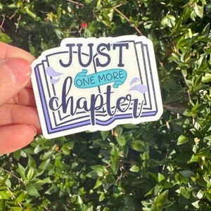 May include: A white sticker with the text "JUST ONE MORE chapter" in a stylized font. The sticker has a book design with purple and teal accents. The sticker is held up against a background of green leaves.