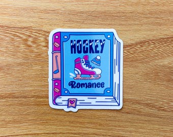 Może przedstawiać: Naklejka z motywem książki z napisem "HOCKEY Romance" w kolorze niebieskim i różowym. Okładka ma jasnoniebieskie tło z różową łyżwą i zimową czapką. Naklejka ma białą obwódkę.