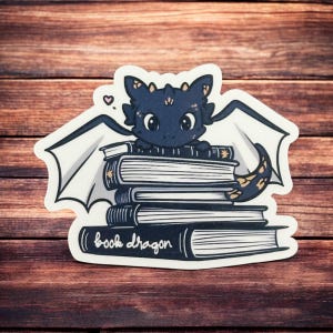 May include: A black and blue book dragon with gold accents sits on top of a stack of books. The books have white pages and black covers. The text "book dragon" is written on the bottom book.