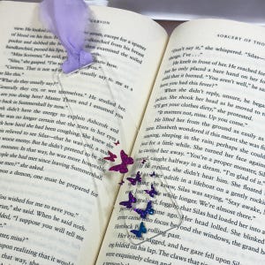 May include: A close-up of a book with a purple and pink butterfly bookmark. The book is open to a page with the text "Sorcery of the" and "Don't say it," she whispered. "Silas-things. I've..."