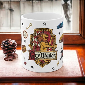 May include: White ceramic mug featuring the Gryffindor house crest from the Harry Potter series. The crest includes a lion, the house name, and other themed illustrations. The mug is decorated with stars, a key, and a lightning bolt.