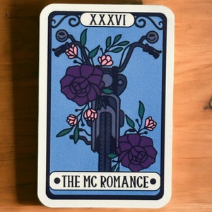 May include: A tarot card illustration depicting a motorcycle handlebar decorated with purple roses and pink flowers. The card has a blue backdrop and the text "THE MC ROMANCE" at the bottom. The top of the card reads "XXXVI".
