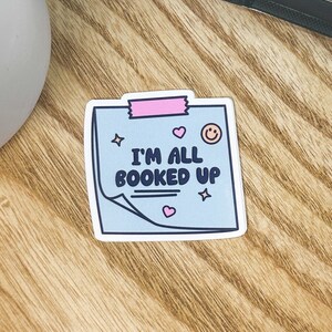 Puede incluir: Una pegatina azul y blanca con cinta rosa y una cara sonriente. La pegatina dice "I'm all booked up" con un corazón rosa y una estrella a cada lado.