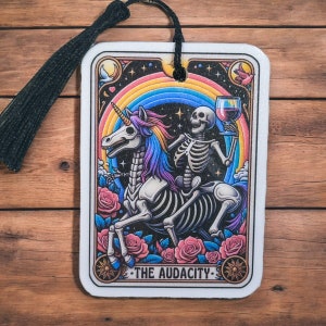 May include: A white card with a black border and a colorful illustration of a skeleton riding a unicorn. The unicorn is rainbow colored and the skeleton is holding a glass of red wine. The card has the text "THE AUDACITY" at the bottom.