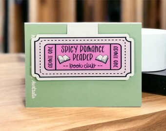 Könnte beinhalten: Ein rosa und schwarzer Aufkleber in Ticketform mit dem Text "SPICY ROMANCE READER book club" und zwei Buchillustrationen. Der Aufkleber ist auf einer hellgrünen Karte angebracht, mit weißem Hintergrund und Holzoberfläche.
