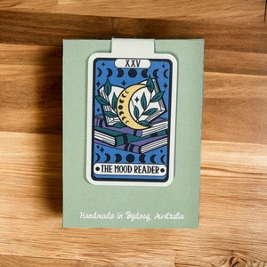 Könnte beinhalten: Eine grüne Karte mit einer blauen und weißen Illustration eines Halbmondes und gestapelter Bücher. Die Karte trägt den Text "XXV" und "THE MOOD READER". Die Karte ist handgefertigt in Sydney, Australien.