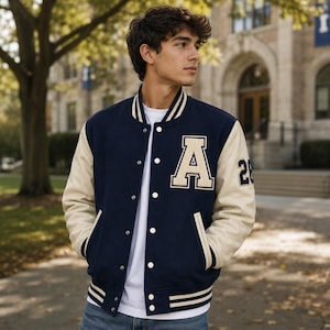 May include: A navy blue varsity jacket with cream-colored sleeves, featuring a large cream letter A on the left chest, the number 26 on the left sleeve, a striped ribbed collar, cuffs, and hem, and silver snap buttons over a white t-shirt.