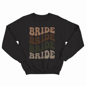 May include: Black sweatshirt with the word "BRIDE" repeated four times in a retro font. Each word is a different shade of brown, tan, and green.
