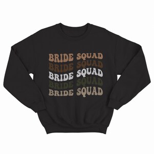 May include: Black sweatshirt with the text "BRIDE SQUAD" repeated five times in a retro font. Each word is a different shade of brown, beige, and green.