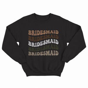 May include: Black sweatshirt with the word "BRIDESMAID" repeated five times in a retro font, with each word in a different shade of brown, green, and white.