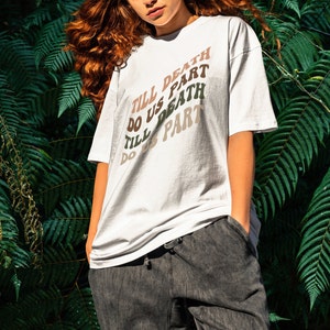 May include: A white t-shirt with the text "Till Death Do Us Part" repeated four times in a rainbow gradient design. The shirt is worn by a person with long, curly red hair.