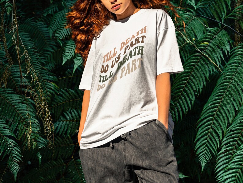 May include: A white t-shirt with the text "Till Death Do Us Part" repeated four times in a rainbow gradient design. The shirt is worn by a person with long, curly red hair.
