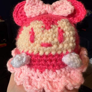 May include: A handmade crochet doll in shades of pink and cream. The doll features a bow, ears, and a ruffled skirt. The face has embroidered details. The doll is a cute, whimsical design.