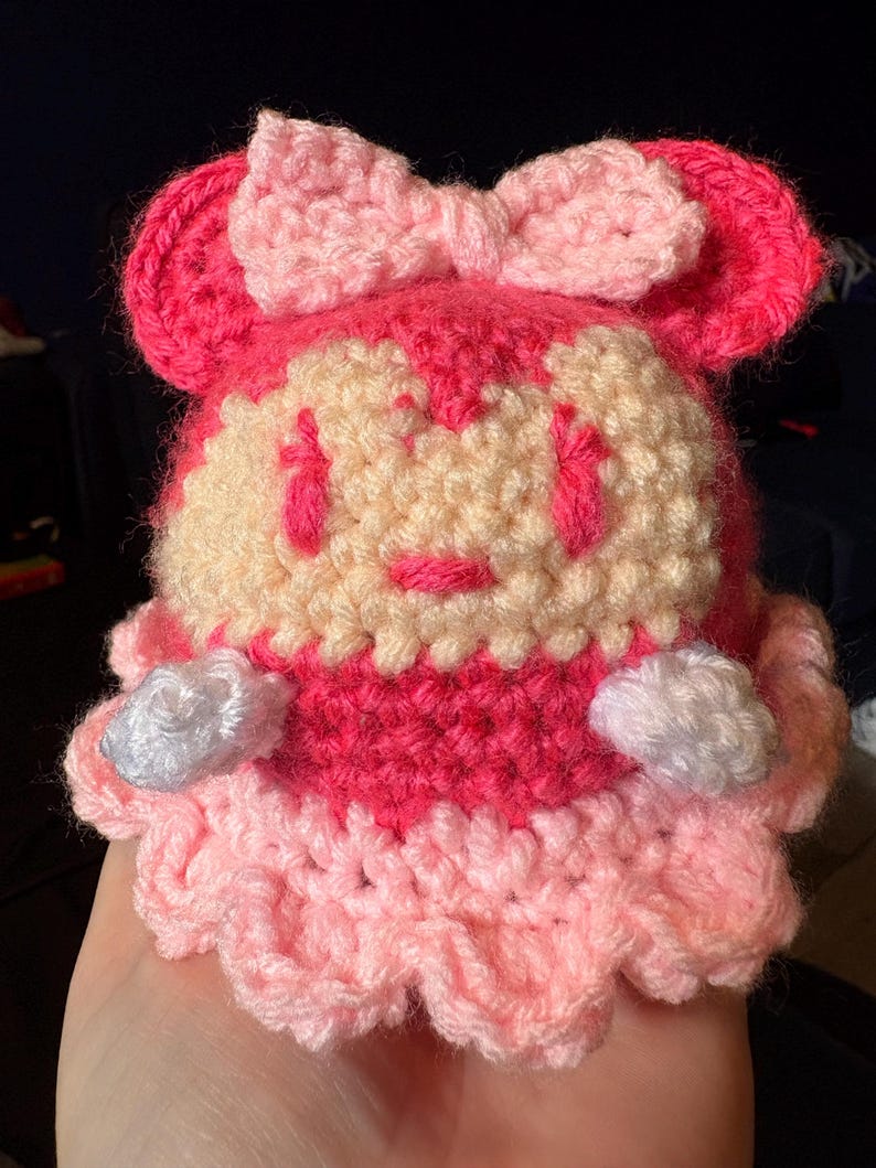 May include: A handmade crochet doll in shades of pink and cream. The doll features a bow, ears, and a ruffled skirt. The face has embroidered details. The doll is a cute, whimsical design.