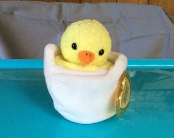 Peut inclure: Un poussin en peluche jaune dans un récipient en peluche blanc en forme d'œuf. Le poussin a des yeux noirs et un bec orange. Le récipient a une étiquette dorée attachée.