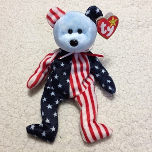 May include: A blue and white beanie baby bear wearing a red, white, and blue outfit with stars and stripes. The bear has a red and white Ty tag attached to its ear.