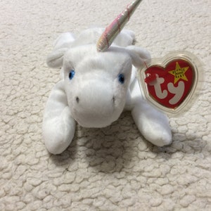 Op de afbeelding: Witte pluche eenhoorn met een regenbooghoorn en blauwe ogen. De eenhoorn ligt met zijn hoofd naar de zijkant gedraaid. Hij heeft een Ty Beanie Babies-label aan zijn been.