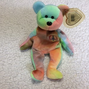 May include: A tie-dye plush bear with a peace sign on its chest. The bear is a light blue, green, orange, and yellow color. The bear has black eyes and a black nose.