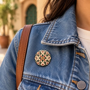 May include: A circular enamel pin featuring a symmetrical mandala-style pattern in shades of deep green, red, and cream. The design includes intricate floral-inspired motifs and pointed petal shapes arranged around a central star-like point.