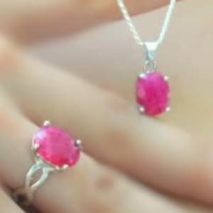 以下が含まれることがあります： オーバルカットの鮮やかなピンク色の宝石をあしらった、シルバーのネックレスとリングのセット。ペンダントは繊細なチェーンから吊り下げられ、リングはツイストバンドのデザインです。ジュエリーは人の手で展示されています。
