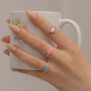 Puede incluir: Tres anillos de oro con piedras de ópalo de forma ovalada. Los anillos están en la mano de una persona, con piedras blancas, rosas y azules.