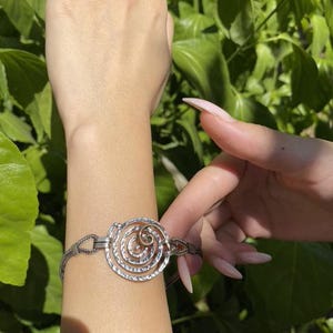 May include: A silver-toned bracelet with a spiral design. The bracelet features a central circular element with a textured spiral pattern, connected by a chain-like band. The bracelet is worn on a wrist.