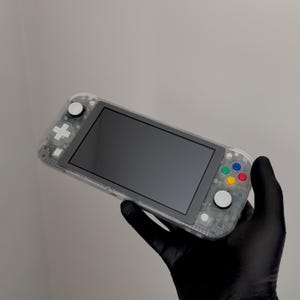 Könnte beinhalten: Eine tragbare Spielkonsole mit einem transparenten Kunststoffgehäuse. Das Gerät verfügt über einen schwarzen Bildschirm, weiße Analogsticks, ein weißes Steuerkreuz und vier Hauptaktionstasten in den Farben Blau, Grün, Gelb und Rot.