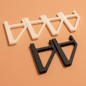 May include: Four white and two black plastic triangular brackets are arranged on a peach-colored background. The brackets have a unique shape, with a flat top and a triangular opening. The black brackets have a textured surface.