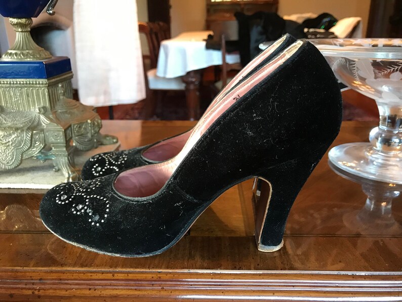 K&ouml;nnte beinhalten: Ein Paar schwarze Samt-High-Heel-Schuhe mit spitzer Zehenpartie und einem dekorativen Design aus silbernen Nieten. Die Schuhe haben ein rosafarbenes Futter und einen hohen Absatz.