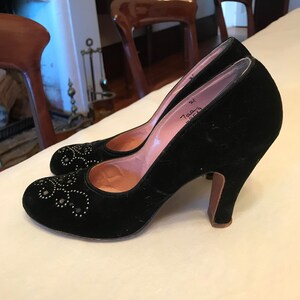 K&ouml;nnte beinhalten: Ein Paar schwarze Pumps aus Samt mit spitzer Zehenpartie und hohem Absatz. Die Schuhe sind mit silberfarbenen Nieten verziert und haben eine rosa Lederinnensohle. Die Schuhe stehen auf einem wei&szlig;en Tisch.