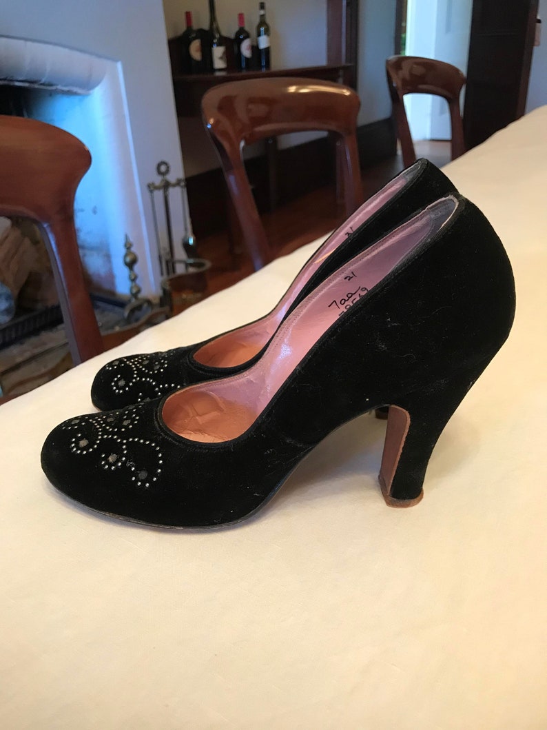 K&ouml;nnte beinhalten: Ein Paar schwarze Pumps aus Samt mit spitzer Zehenpartie und hohem Absatz. Die Schuhe sind mit silberfarbenen Nieten verziert und haben eine rosa Lederinnensohle. Die Schuhe stehen auf einem wei&szlig;en Tisch.