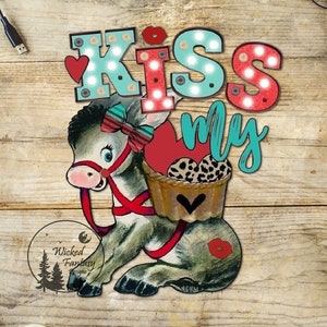 Può includere: Un'illustrazione fantasiosa di un asino che trasporta un cesto con un cuore e un design a stampa leopardata. L'asino indossa un nastro rosso e il testo "Kiss My" è scritto in lettere colorate con lampadine intorno alle lettere.