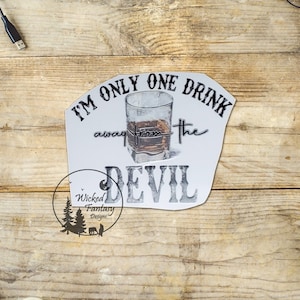 May include: A white sign with the text "I'M ONLY ONE DRINK away from the DEVIL" and a graphic of a whiskey glass. The sign is on a wooden surface. The sign also has the logo for Wicked Fantasy Designs.