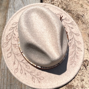 May include: A beige felt fedora hat with a brown leather band and white beads. The hat has a delicate floral design embroidered around the brim.