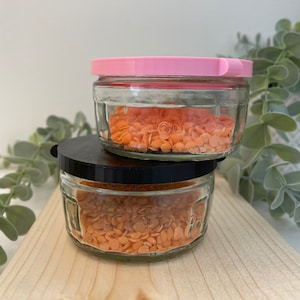 May include: Two clear glass jars with orange contents, one stacked on top of the other. The top jar has a pink lid, and the bottom jar has a black lid. The jars are on a wooden surface, with greenery in the background.