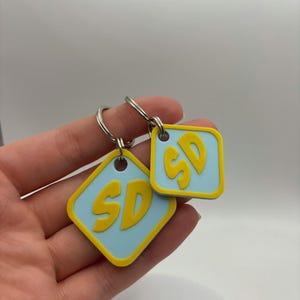 May include: Two keychains with a light blue square background and a yellow diamond outline. The letters "SD" are in yellow. The keychains are attached to silver keyrings. The background is white.