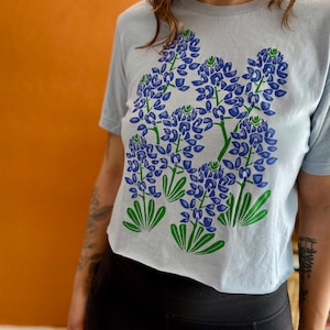 May include: A light blue t-shirt featuring a graphic of bluebonnet flowers in shades of blue and green. The floral design is centered on the front of the shirt. The shirt has short sleeves and a crew neck.
