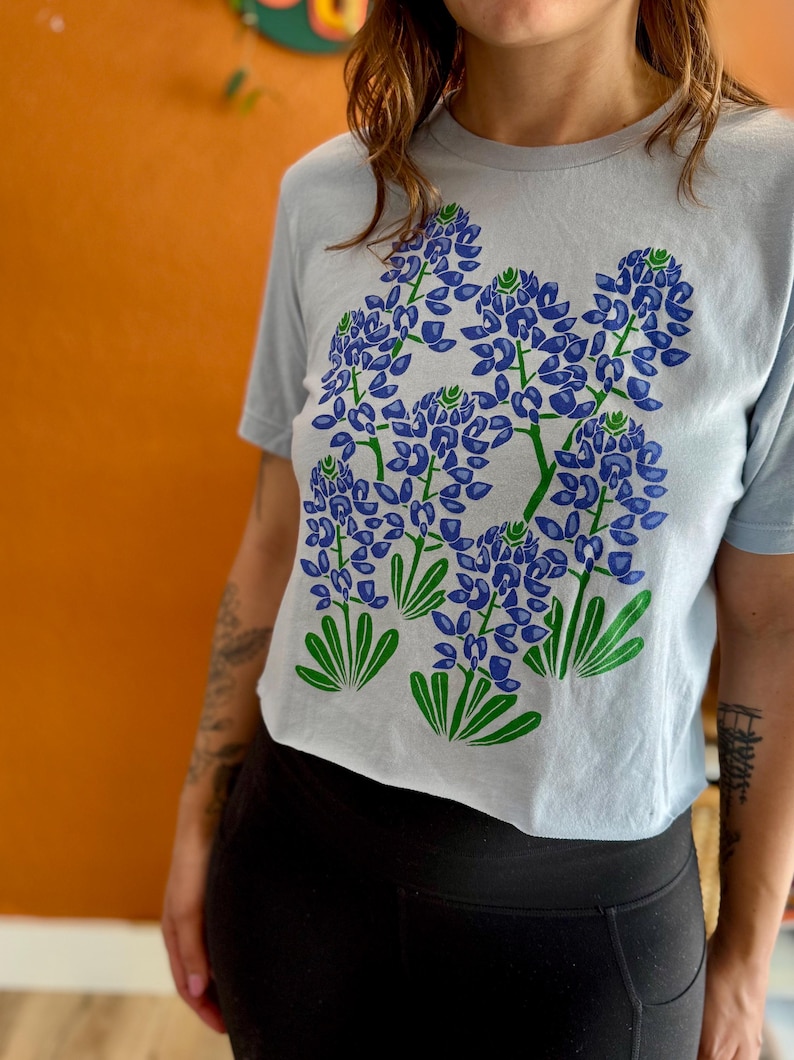May include: A light blue t-shirt featuring a graphic of bluebonnet flowers in shades of blue and green. The floral design is centered on the front of the shirt. The shirt has short sleeves and a crew neck.