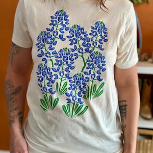 May include: Cream-colored t-shirt featuring a bluebonnet floral design in blue and green. The graphic depicts several clusters of blue flowers with green stems and leaves. The shirt has short sleeves and a crew neck.