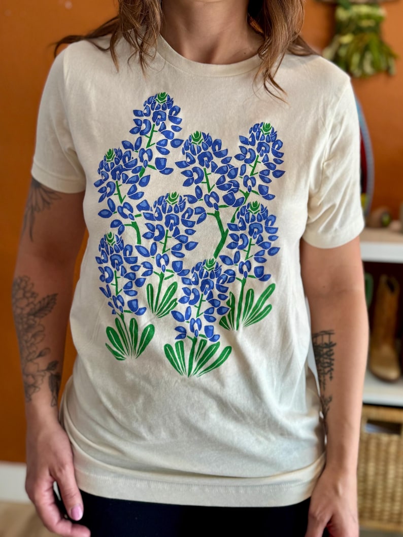 May include: Cream-colored t-shirt featuring a bluebonnet floral design in blue and green. The graphic depicts several clusters of blue flowers with green stems and leaves. The shirt has short sleeves and a crew neck.