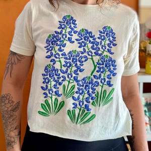 May include: Cream-colored t-shirt featuring a large bluebonnet floral design in blue and green. The short-sleeved shirt has a crew neck and a cropped fit. The graphic depicts several clusters of blue flowers with green leaves.
