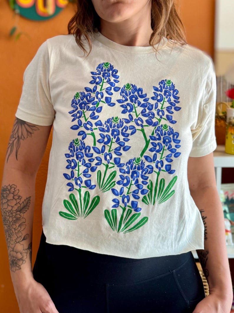 May include: Cream-colored t-shirt featuring a large bluebonnet floral design in blue and green. The short-sleeved shirt has a crew neck and a cropped fit. The graphic depicts several clusters of blue flowers with green leaves.