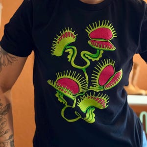 May include: Black t-shirt featuring a vibrant graphic of Venus flytraps. The design showcases multiple open traps in shades of red and green, with detailed serrated edges and stems. A botanical-themed apparel item.