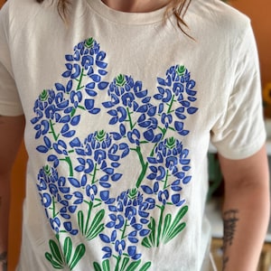 以下が含まれることがあります： 青い花と緑の葉のテキサスブルーボネットのプリントが施された白いTシャツです。